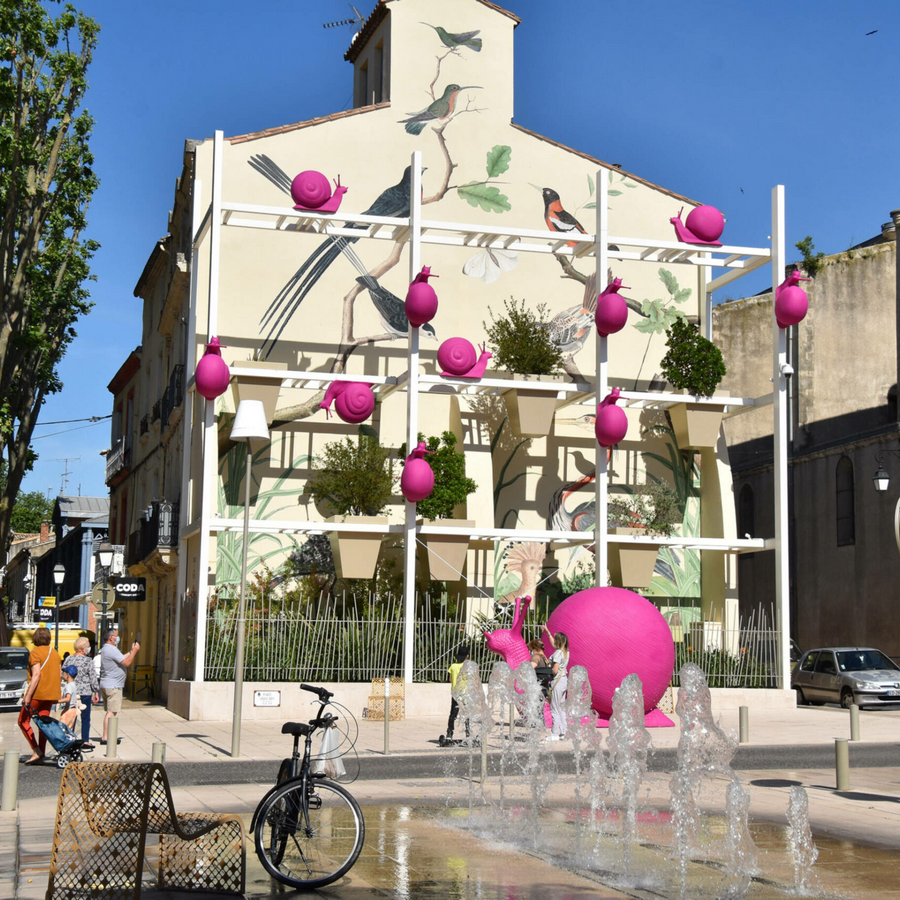 Façade décorée d’une fresque d’oiseaux avec installations roses et jets d’eau sur une place à Lunel